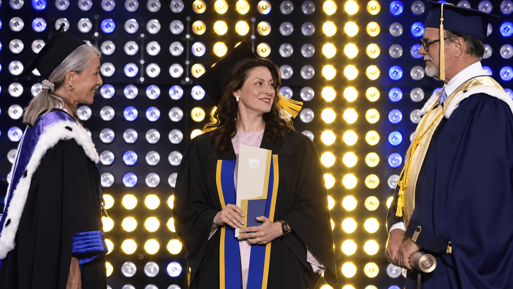 Diplômée souriante tenant son diplôme entre deux officiels académiques devant un mur de lumières.